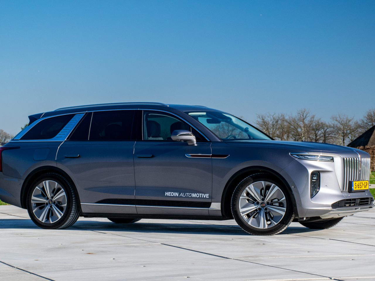 Buiten staat een grijze Hongqi E-HS9 SUV geparkeerd op een betonnen oppervlak, met het opschrift 'Hedin Automotive'. Het voertuig heeft een strak ontwerp met grote wielen en een moderne grille.