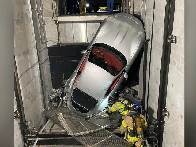 Een zilveren Ferrari zit schuin in een liftschacht terwijl brandweerlieden de situatie onderzoeken. Het puin ligt verspreid over de onderkant van de auto.