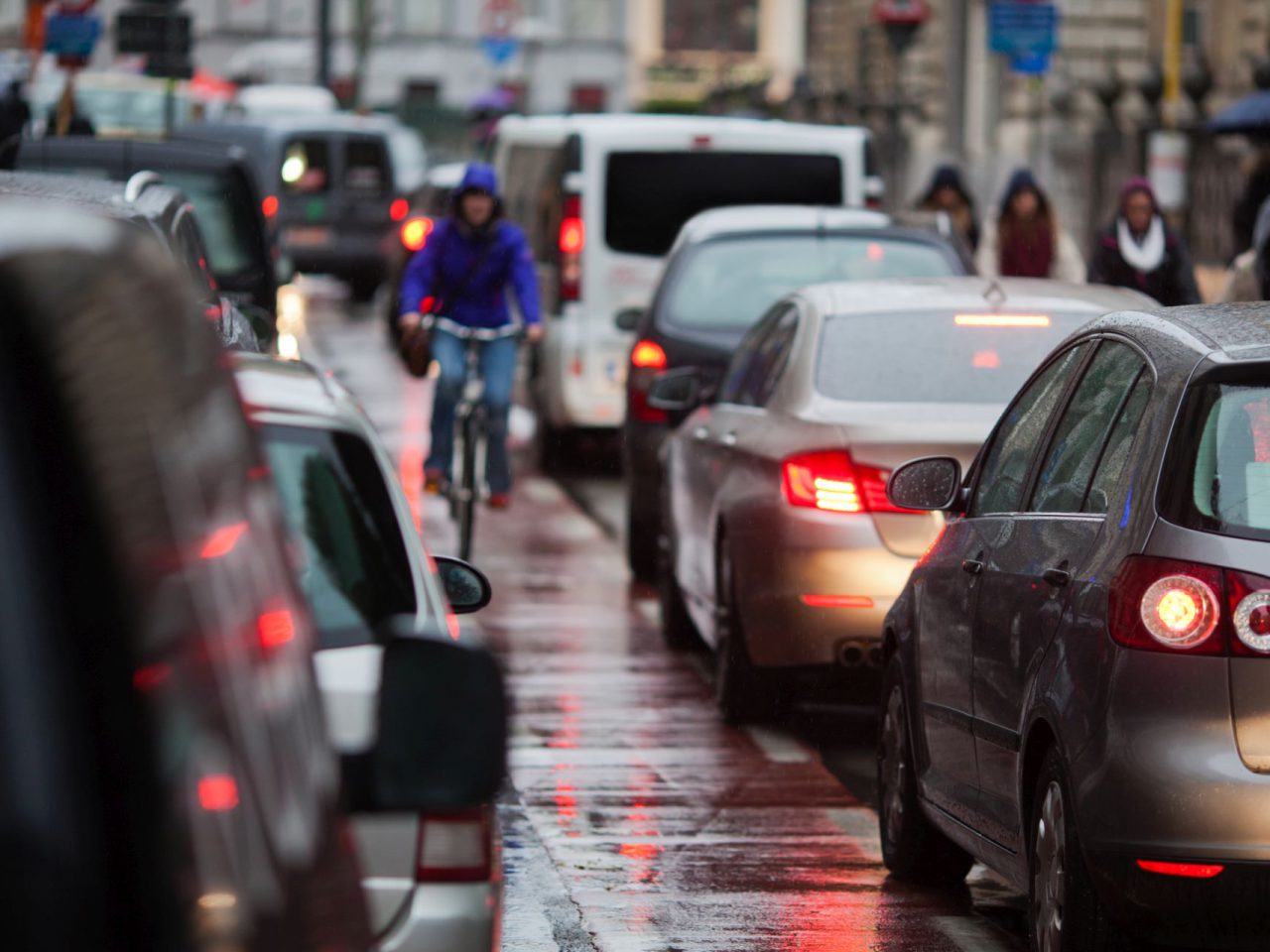 Stadsstraat met zwaar verkeer op een regenachtige dag; auto's opgesteld en een fietser in een blauwe jas die tussen voertuigen navigeert, waarbij een van de drukste filesteden van Nederland wordt getoond.