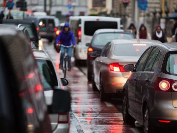 Stadsstraat met zwaar verkeer op een regenachtige dag; auto's opgesteld en een fietser in een blauwe jas die tussen voertuigen navigeert, waarbij een van de drukste filesteden van Nederland wordt getoond.