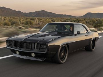 Een zwarte klassieke Camaro-musclecar rijdt bij zonsondergang langs een weg met bergen en woestijnlandschap op de achtergrond.