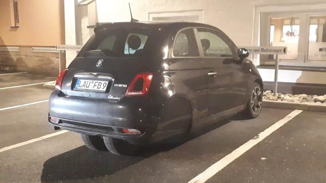 Een zwarte Fiat 500 Hybrid Ellenator staat geparkeerd op een aangewezen parkeerplaats op een buitenterrein. Het voertuig, met kenteken "LAU-FB9", valt op tussen de omringende voertuigen.