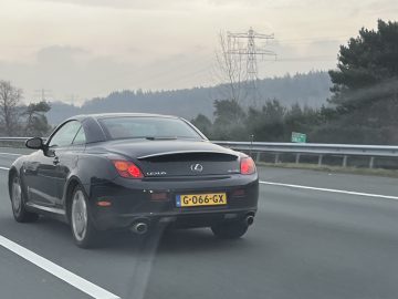 Een zwarte Lexus SC430 met een geel kenteken rijdt op een snelweg omzoomd door bomen en hoogspanningsleidingen onder een bewolkte hemel.