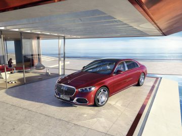 Een luxe rode Maybach sedan staat geparkeerd op het terras van een modern kusthuis met glazen wanden en uitzicht op de oceaan.