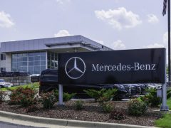 Voor een strak gebouw met glazen gevel staat een Mercedes-Benz-dealer met een prominent logobord. Het perceel, badend in het zonlicht, is gevuld met een reeks ongerepte Mercedes-voertuigen.