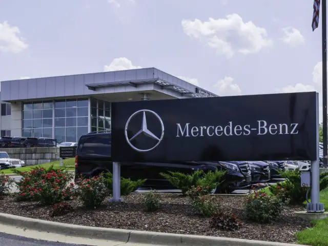 Voor een strak gebouw met glazen gevel staat een Mercedes-Benz-dealer met een prominent logobord. Het perceel, badend in het zonlicht, is gevuld met een reeks ongerepte Mercedes-voertuigen.