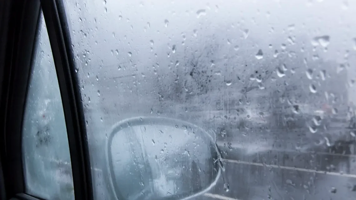 Regendruppels op een mistige autoruit met beslagen ruiten belemmeren gedeeltelijk het zicht op de zijspiegel en een bewolkt tafereel buiten.