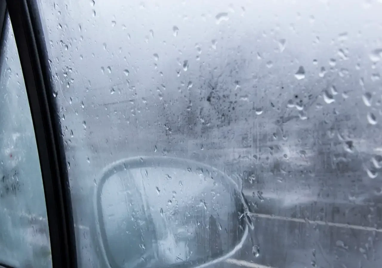 Regendruppels op een mistige autoruit met beslagen ruiten belemmeren gedeeltelijk het zicht op de zijspiegel en een bewolkt tafereel buiten.