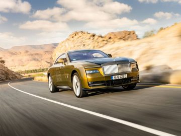 Een gouden Rolls-Royce Spectre glijdt over een kronkelige bergweg onder een gedeeltelijk bewolkte hemel.