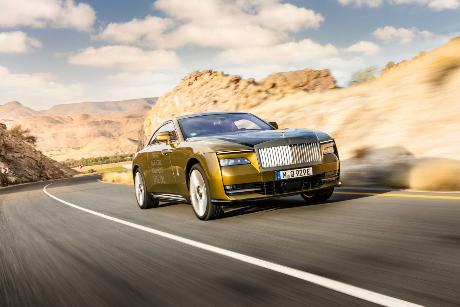 Een gouden Rolls-Royce Spectre glijdt over een kronkelige bergweg onder een gedeeltelijk bewolkte hemel.