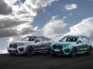 Twee moderne BMW SUV's, een zilveren en een groene BMW X5 M Competition, staan geparkeerd op een verharde ondergrond onder een gedeeltelijk bewolkte hemel.