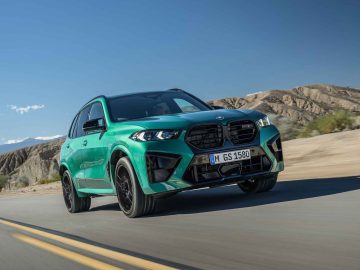 Een groene BMW X5 M Competition met BMW-embleem rijdt over een woestijnweg onder een helderblauwe lucht, bergen op de achtergrond. Op het kenteken staat GM GS 1580.