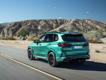 Een groene BMW X5 M Competition rijdt op een verharde weg in een woestijnlandschap met rotsachtige heuvels op de achtergrond onder een helderblauwe lucht.