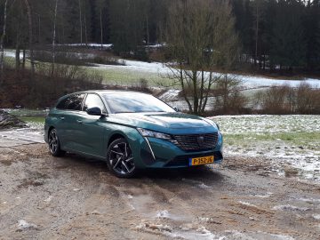 Een groene Peugeot 308 SW staat geparkeerd op een modderige en gedeeltelijk besneeuwde grond voor een bosrijk gebied. Op het kenteken staat "P-352-JG".