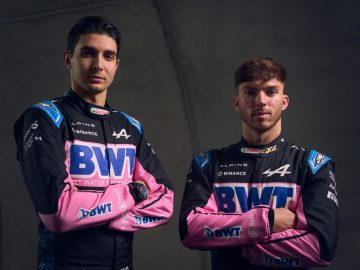 Twee coureurs, gekleed in Alpine F1 Team-pakken met BWT-sponsorlogo's, staan zij aan zij in een professionele studioomgeving, wat de geest van Alpine's concurrentievoordeel belichaamt.