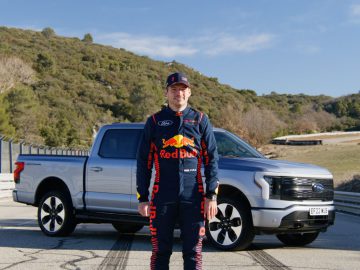 Een persoon in een blauw en rood racepak staat voor een zilveren Ford pick-up op een weg met heuvels en bomen op de achtergrond.
