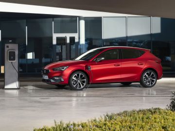 Een rode elektrische auto staat, samen met enkele plug-in hybrides, geparkeerd en wordt opgeladen bij een station voor een modern gebouw.