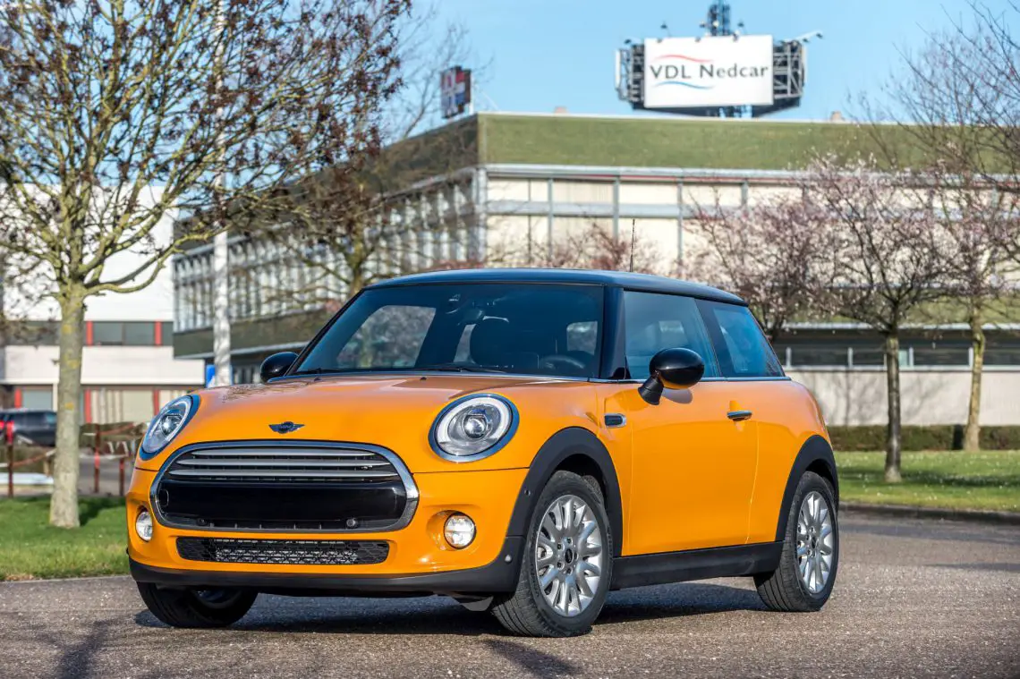 Ein orangefarbener Mini Cooper parkt an einem klaren Tag vor dem Fabrikgebäude von VDL Nedcar, mit Bäumen im Hintergrund.