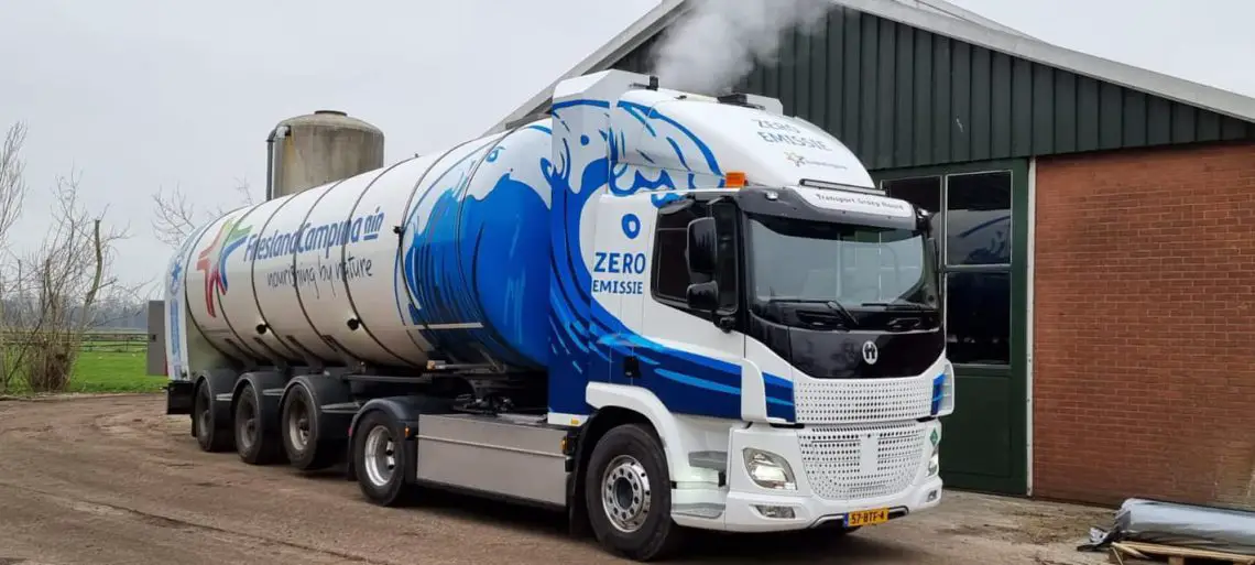 Een grote witte vrachtwagen met blauwe golfafbeeldingen en de tekst "ZERO EMISSIE" staat geparkeerd bij een gebouw met een schoorsteen die stoom of rook uitstraalt. De vrachtwagen, onderdeel van het bedrijf FrieslandCampina, toont haar inzet voor duurzaamheid door te rijden op waterstof.