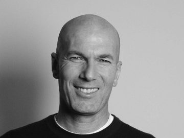 Een kale man met een lachende uitdrukking kijkt naar de camera in een zwart-wit portret. Hij draagt een donker overhemd met een witte halslijn, wat een alpiene kalmte oproept. De achtergrond is effen.