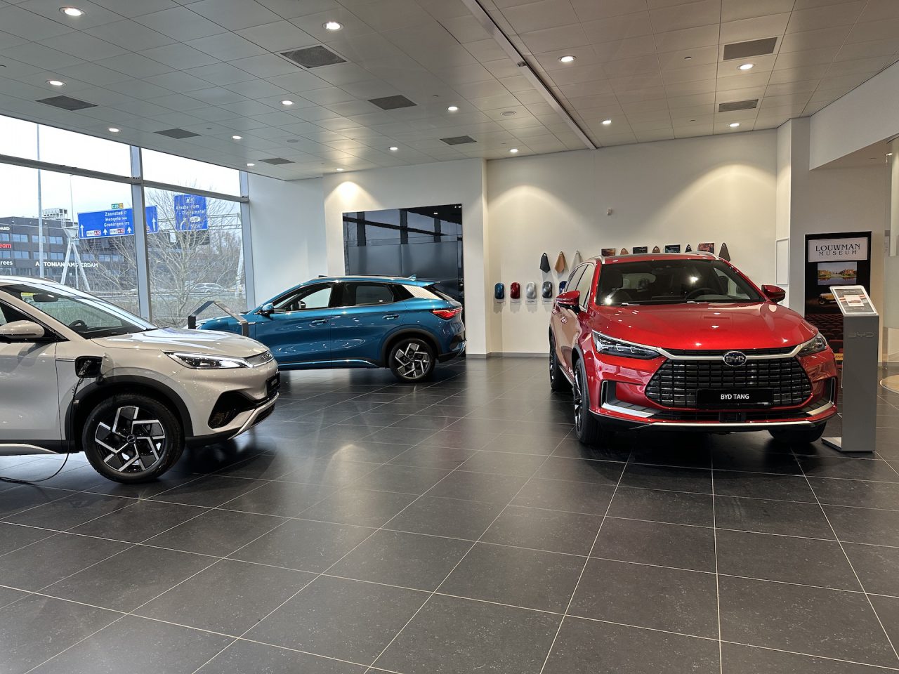 Drie auto's, waaronder de BYD Han, worden tentoongesteld in een moderne showroom met grote ramen. De BYD Han aan de linkerkant is aangesloten op een laadstation, terwijl de andere twee in de kijkstand staan.
