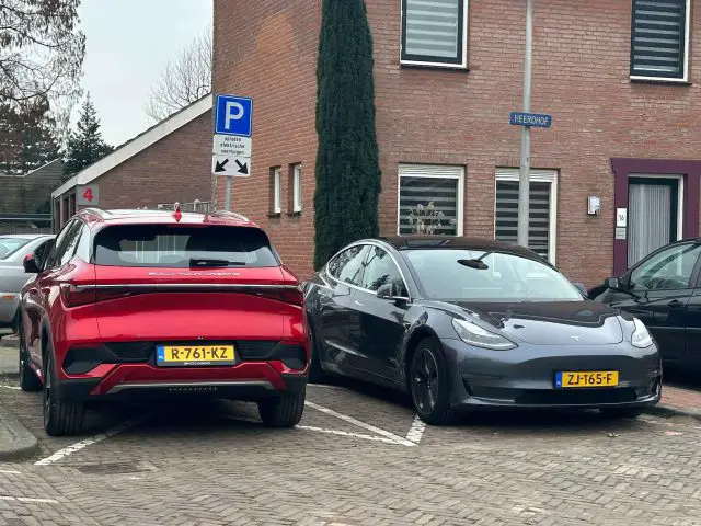 Twee auto's, een rode SUV en een donkergekleurde sedan, staan naast elkaar geparkeerd op aangewezen parkeerplaatsen voor een bakstenen gebouw. Beide auto's hebben gele kentekenplaten en de SUV staat iets over de lijn geparkeerd. De scène ziet eruit alsof deze rechtstreeks uit een BYD Atto 3-recensie kan komen.