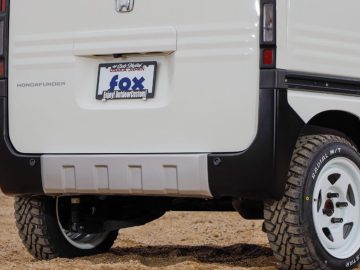 Achteraanzicht van een wit Hondafunder-voertuig met offroad-banden en een kentekenplaat met de tekst 'vos'. Het voertuig lijkt stil te staan op een vuil oppervlak.