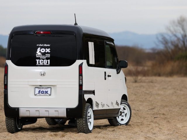 Een wit-zwarte Hondafunder-minibus staat geparkeerd op een zanderig terrein met stickers op de ramen en de carrosserie. Het voertuig staat op witte wielen en op de achtergrond is een heuvelachtig landschap zichtbaar.