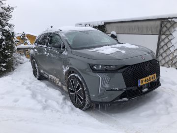 Een grijze DS 7 E-TENSE SUV staat geparkeerd op een besneeuwde oprit, met ijspegels die aan de bumper hangen. Het voertuig heeft een Nederlands kenteken met de tekst "R-468-NX", en sneeuw bedekt de bovenkant en delen van de zijkanten.