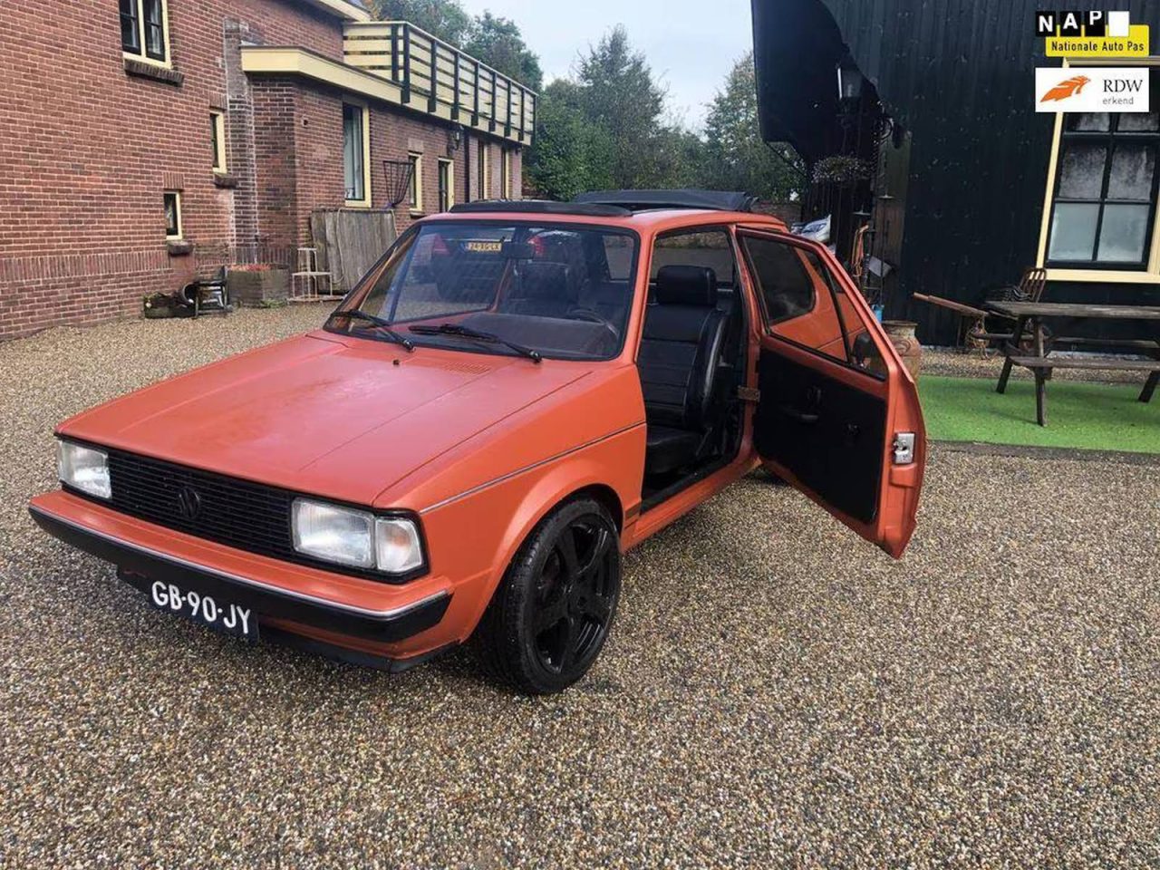 Een vintage oranje Jetta met zwarte velgen staat geparkeerd op een grindoprit voor een gebouw. De bestuurdersdeur staat open en het Nederlandse kenteken GB-90-JY is zichtbaar.