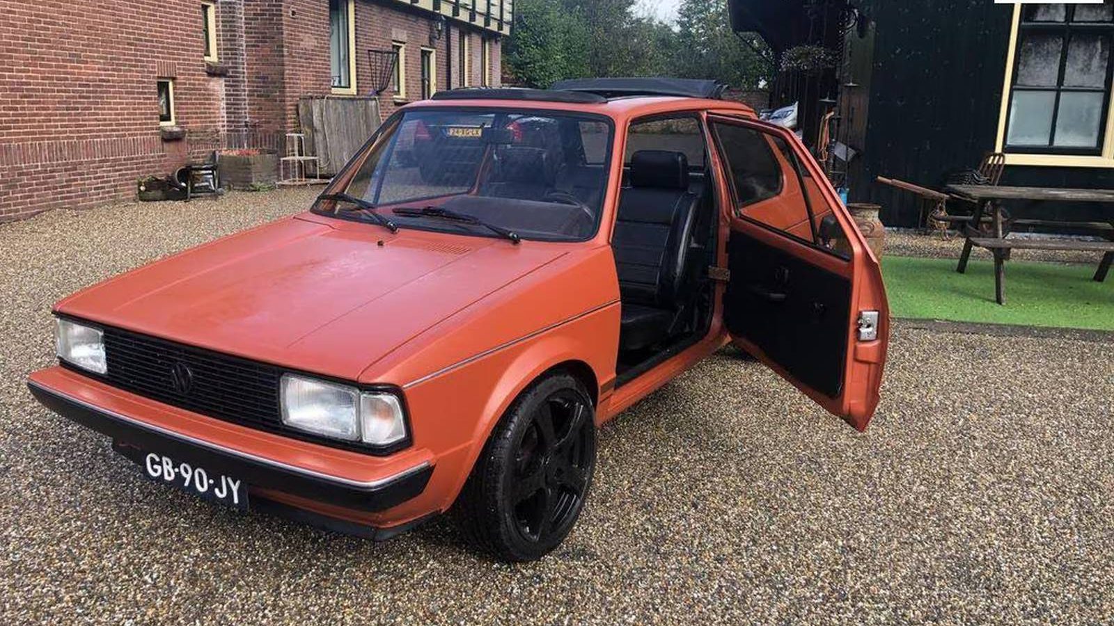 Een vintage oranje Jetta met zwarte velgen staat geparkeerd op een grindoprit voor een gebouw. De bestuurdersdeur staat open en het Nederlandse kenteken GB-90-JY is zichtbaar.