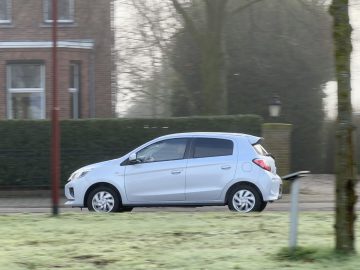 Een zilveren Mitsubishi Space Star glijdt door een straat in een buitenwijk met een heg, een bakstenen gebouw en bomen op de achtergrond. De weg lijkt enigszins bevroren.