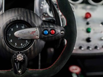 Close-up van een autodashboard met een Pagani Zonda R Evo-logo, een snelheidsmeter, stuurbedieningen met het opschrift "Flash" en "DRS" en een deel van een bedieningspaneel met verschillende schakelaars en knoppen.