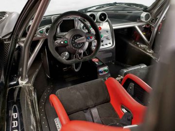 Close-up van een Pagani Zonda R Evo-interieur met een stuur met meerdere bedieningselementen, een gedetailleerd dashboard met verschillende meters en knoppen, en zwarte en rode kuipstoelen.