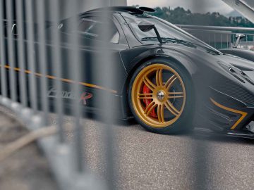 Een close-up van een zwarte Pagani Zonda R Evo met gouden wielen, gezien door metalen balustrades. De auto lijkt geparkeerd op een verhard oppervlak met wazig groen op de achtergrond.