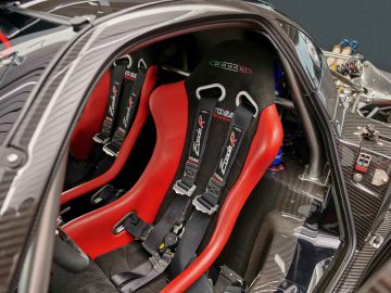 Close-up van een auto-interieur met rode racestoelen met harnassen en een rolkooi, die doet denken aan de Pagani Zonda R Evo. De stoelen zijn voorzien van "Cobra" -logo's en de auto is voorzien van een koolstofvezelstructuur.