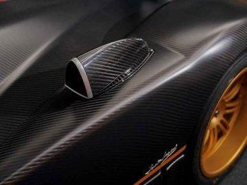 Close-up van het koolstofvezel exterieur en de zijspiegel van een Pagani Zonda R Evo, met een aerodynamisch ontwerp met een zwarte afwerking en goudkleurige wielen zichtbaar op de achtergrond.