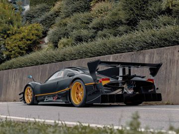 Een strakke zwarte Pagani Zonda R Evo met gele velgen en een opvallende achtervleugel staat geparkeerd op een weg naast een betonnen muur en groene struiken.