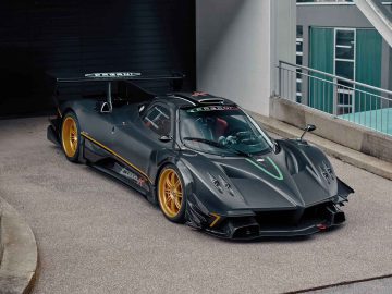 Een zwarte Pagani Zonda R Evo met gele velgen staat geparkeerd op een betonnen ondergrond bij een gebouw. De auto heeft een gestroomlijnd ontwerp met een grote achtervleugel en aerodynamische kenmerken.