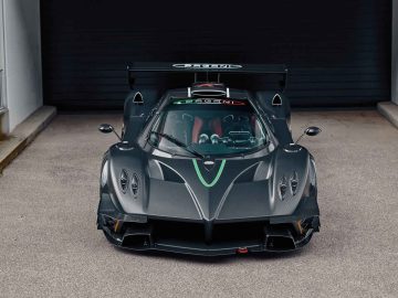 Een Pagani Zonda R Evo, voorzien van een zwart exterieur en een grote achterspoiler, staat geparkeerd voor een grijze garagedeur. De auto heeft een strak design met groene en rode accenten.