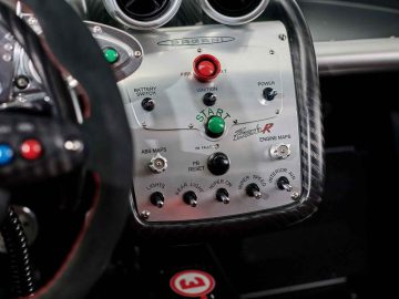 Close-up van het bedieningspaneel van een Pagani Zonda R Evo met knoppen voor contact, vuur, stroom, ruitenwisser, licht, ABS en meer, met de merknaam "Pagani" prominent weergegeven.