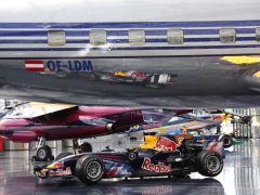 Een Formule 1-auto van Red Bull Racing, bestuurd door de Nederlandse Max Verstappen, wordt binnenshuis tentoongesteld onder een vliegtuig, met een ander vliegtuig zichtbaar op de achtergrond.