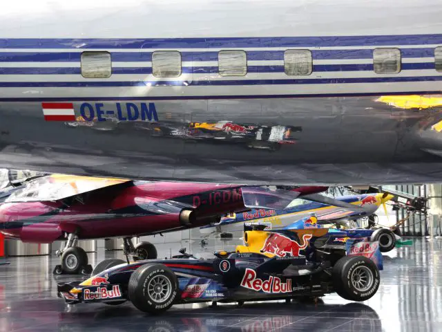Een Formule 1-auto van Red Bull Racing, bestuurd door de Nederlandse Max Verstappen, wordt binnenshuis tentoongesteld onder een vliegtuig, met een ander vliegtuig zichtbaar op de achtergrond.