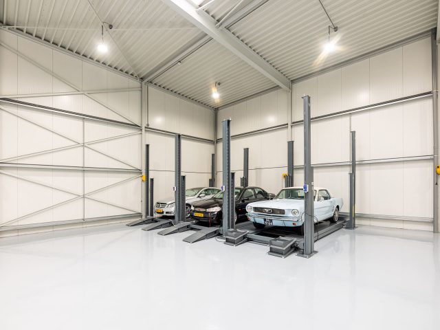 Drie klassieke auto's staan geparkeerd op autoliften in een strakke, goed verlichte industriële garage met witte muren en een metalen structuur, die doet denken aan de nauwgezette ontwerpstijl van Rick van Stippent.