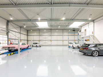 Een ruime, goed verlichte garage met vier auto's, waaronder één op een hydraulische lift van Rick van Stippent, met gepolijste witte vloeren en smetteloos witte muren.