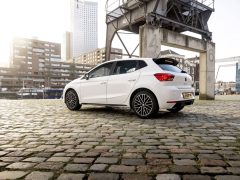 Witte Ibiza hatchback-auto geparkeerd op een geplaveide straat met industriële en stedelijke gebouwen op de achtergrond.