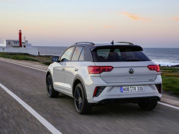 Een witte Volkswagen T-Roc navigeert sierlijk over een kustweg, met een vuurtoren en de oceaan als schilderachtige achtergrond tijdens een adembenemende zonsondergang.