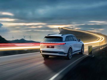 Een witte XPENG G9 SUV rijdt in de schemering over een bochtige weg, met windturbines op de achtergrond en lichtsporen die beweging aangeven op het gebogen gedeelte verderop.