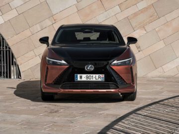 Vooraanzicht van een Lexus RZ met een bruin en zwarte buitenkant, geparkeerd op een geplaveid terrein tegen een beige stenen muur.