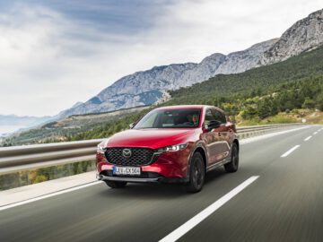 Een rode Mazda SUV die over een snelweg door een bergachtig landschap rijdt onder een gedeeltelijk bewolkte hemel, getuigt van kracht en elegantie.